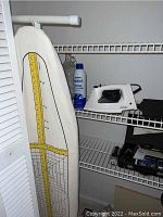 Ironing board with measuring tape design on cloth cover, two irons and spray starch bottle on storage shelving