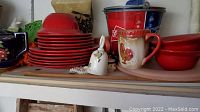 View of Pier One red dishes stacked including plates and bowls, a rooster design ceramic mug, white decorative bell, and Budweiser tin bucket on a table.