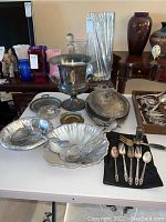 Full view of multiple silver plated bowls, plates, serving utensils, a large urn-style container, serving tray with lid, and brass change tray on a table with other background objects.