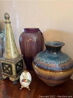 Photo of four lot items: tall faux marble decorative piece on left, purple glass vase in center back, multicolored glazed ceramic vase in front right, hand-painted decorative egg on stand in front left.