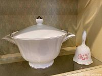 Covered white porcelain serving dish with lid and silver trim next to white ceramic bell with red lettering.