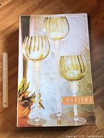 Box front showing three amber glass hurricanes in different sizes with gold flakes, tall slender stems, and ribbed amber tops.