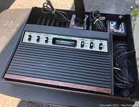 Top-down photo showing the Atari 2600 console in a black plastic organizer tray along with two joysticks and cables, placed on a textured surface in outdoor lighting.