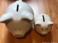 Front view of large and small white ceramic piggy banks showing coin slots and facial features