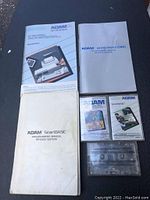 Photo showing four Coleco ADAM computer manuals and two cassette tapes, along with a clear cassette case.
