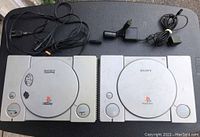 Front top view of two Sony PlayStation 1 consoles, showing power and open buttons, disc lids, cables attached or nearby.