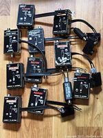 Top-down view showing 11 black vintage game switches labeled mainly Coleco Adam with cables and connectors assorted on wooden floor surface.