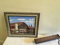 Framed oil painting of barn and silo resting on a white textured cloth with a small dark wooden shelf placed beside it.
