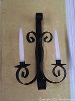 Photo of one black metal wall sconce mounted on wall holding two white taper candles, showing scroll design and drip cup holders.
