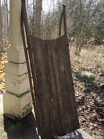 Full view of antique wooden sleigh standing upright outdoors, showing deck, side rails and overall shape.