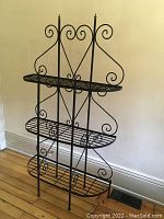 Three shelf black metal baker's stand with scroll details standing on wooden floor against beige wall, showing full front view.