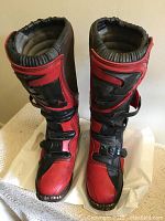 Front view of red and black dirt bike boots standing upright on white surface showing padded collar, straps, and textured sole.