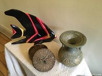 Photo showing the fish ornament in black with red and yellow accents, the solid urn with carved detailing, and the round woven basket with lid on a table.