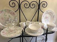 Photo showing the full set of floral patterned plates, bowls, cups arranged on a black metal stand. The set includes stacked plates, bowls, cups, and mugs all displaying a pink and green floral pattern on white background.