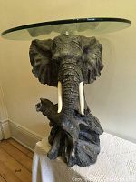 Full view of clear oval glass table top on detailed elephant head base with visible tusks and textured elephant skin details.