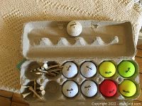 Photo showing golf balls of different brands and colors arranged in an open egg carton style golf ball holder next to assorted wooden golf tees on a beige textured surface.