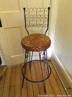 Front view of the metal stool showing the decorative backrest and padded seat on a wooden floor.