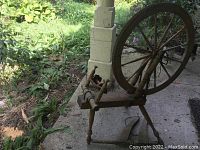 Photo of an older wooden spinning wheel showing the main circular wheel, wooden spindles, and base, placed outside near a concrete pillar with no additional components visible.