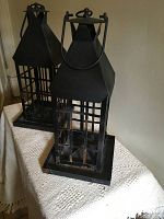 Two older black metal lantern frames showing rust and wear, placed on a textured surface against a plain wall.