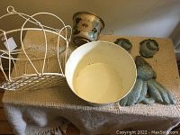 Overall photo showing white metal hanging plant hanger with removable cream-colored pot inside, small round metal plant pot with floral design, and parts of a greenish metal tree face decoration including nose and eye pieces.