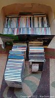 Overview photo showing multiple stacks of music CDs in jewel cases inside a cardboard box and on the floor.