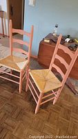 Pair of wooden ladder-back chairs painted in light coral with woven wicker seats shown side by side.