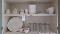 Shelf with white ceramic dishware including plates, bowls, coffee cups, and tea set items. Also includes clear drinking glasses.
