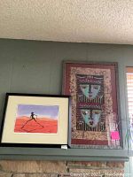 View of both framed artworks on a shelf against a gray wall. The smaller painting on left and larger framed fabric art on right.