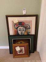 Three framed art prints leaning against a wall, showing varied sizes and subject matter including an abstract face portrait and other scenes.