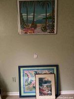 Photo showing one framed tropical coastal scene print hanging on the wall and two prints stacked below it on the floor.