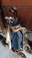 Photo showing the AJAY golf bag filled with twelve golf clubs and the DP golf cart positioned behind it.