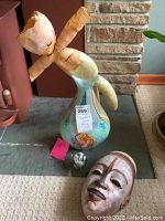 Photo showing handblown glass vase with visible repair at the top, paperweight, paper mache doll wrapped in paper, and wooden carved mask on carpeted floor.
