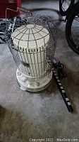 Front view of the white cylindrical kerosene heater with wire cage and control knobs, alongside a black power strip on the floor.