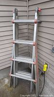 Full front view of the folded Little Giant ladder showing aluminum frame and rungs, red plastic end caps, placed against building siding.
