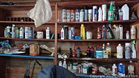 Wide view of multiple shelves showing assorted cleaning products, paint cans, and bottles including liquid plant food, Flex Seal, and paint thinners.