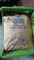 A brown paper bag labeled Cargill North Pacific Boat & Shore Salt, placed inside a green plastic crate, showing boat and globe illustration with blue print.