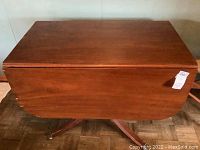 Wooden drop leaf table shown with leaves down, view of tabletop and side with label attached.
