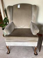 Front view of first beige velvet upholstered wingback arm chair showing padded arms, high back with wings, cushioned seat, and wooden cabriole legs.