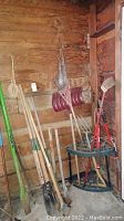 Full view of assorted yard tools leaning against a wooden wall including rakes, shovels, broom, and hand tools organized in a corner.