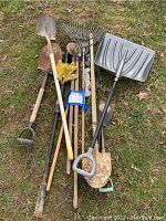 Outdoor tools laid out on grass showing a variety of rakes, shovels, a snow shovel, work gloves, and safety glasses.