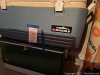 Two large coolers stacked in a basement area. The top cooler is a beige and blue Coleman MaxCold cooler with visible labels and minor wear. The bottom cooler is a dark green Coleman cooler with a latch on the front. Both need cleaning.