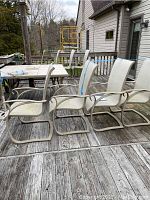 Four beige metal frame patio chairs with mesh seats and backs arranged on wooden deck, showing weathered condition and dirt.