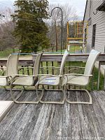 Four beige metal patio chairs with mesh seating arranged on a wooden deck, showing weathered condition and outdoor wear.
