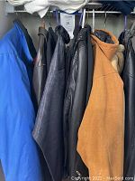 Photo showing six coats hanging, including a blue synthetic coat, leather coats in black and brown, and a tan/beige coat with possible faux fur.