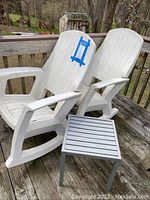 Two white plastic rocking chairs placed next to each other on a wooden deck, alongside a small grey metal table.