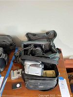 Lot contents showing Canon professional SLR camera in gray carrying bag, Hitachi DVD camcorder in black Hitachi bag, small Canon point-and-shoot camera, and accessories on table.