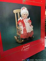 Red box showing an animated Mrs. Claus doll in detailed red and white dress sitting in a wooden rocking chair