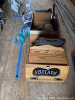 Photo showing wooden bread box with hinged lid and cast iron griddle pan, boxes containing various kitchen items including plates and Pyrex bowls, and blue plastic lids