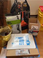 Photo showing first aid kit in white metal case labeled 'FIRST AID KIT 50', knee brace package, organizer backpack and other items in background.