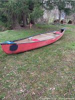 Full side view of the Mohawk canoe showing entire length and red color with black patch at the stern. Canoe is placed on grass outdoors near trees.